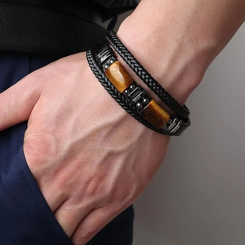 Bracelet pour homme en cuir noir avec pierre œil de tigre, porté sur le poignet gauche. Ce bracelet au design moderne et élégant présente un double cordon en cuir tressé et lisse, associé à un fermoir magnétique, pour un style masculin raffiné.