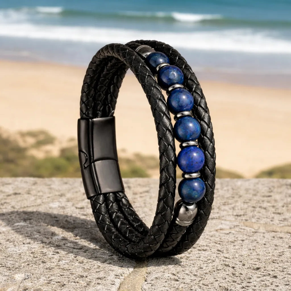 Bracelet Azur en équilibre sur un parapet de pierre face à la mer, soulignant le contraste entre le cuir noir tressé et le bleu profond des six perles de lapis-lazuli.