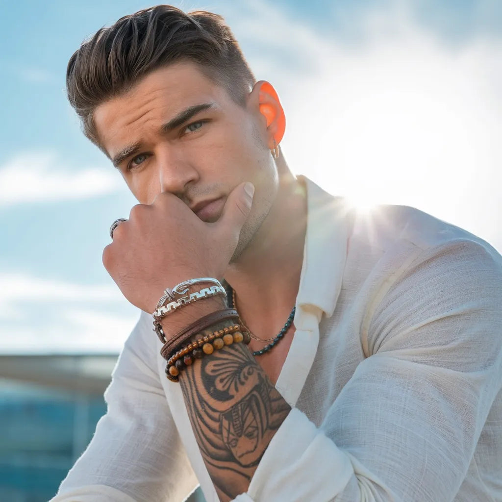 Homme élégant portant une chemise blanche légère, arborant des bracelets multirangs en cuir, perles naturelles et acier argenté. Son bras tatoué ajoute une touche artistique à son style bohème chic, parfait pour les amateurs de mode contemporaine. Capturé sous une lumière naturelle avec un ciel dégagé, cette image évoque liberté et raffinement, mettant en avant l’accessoire comme élément clé d’une tenue moderne et tendance.