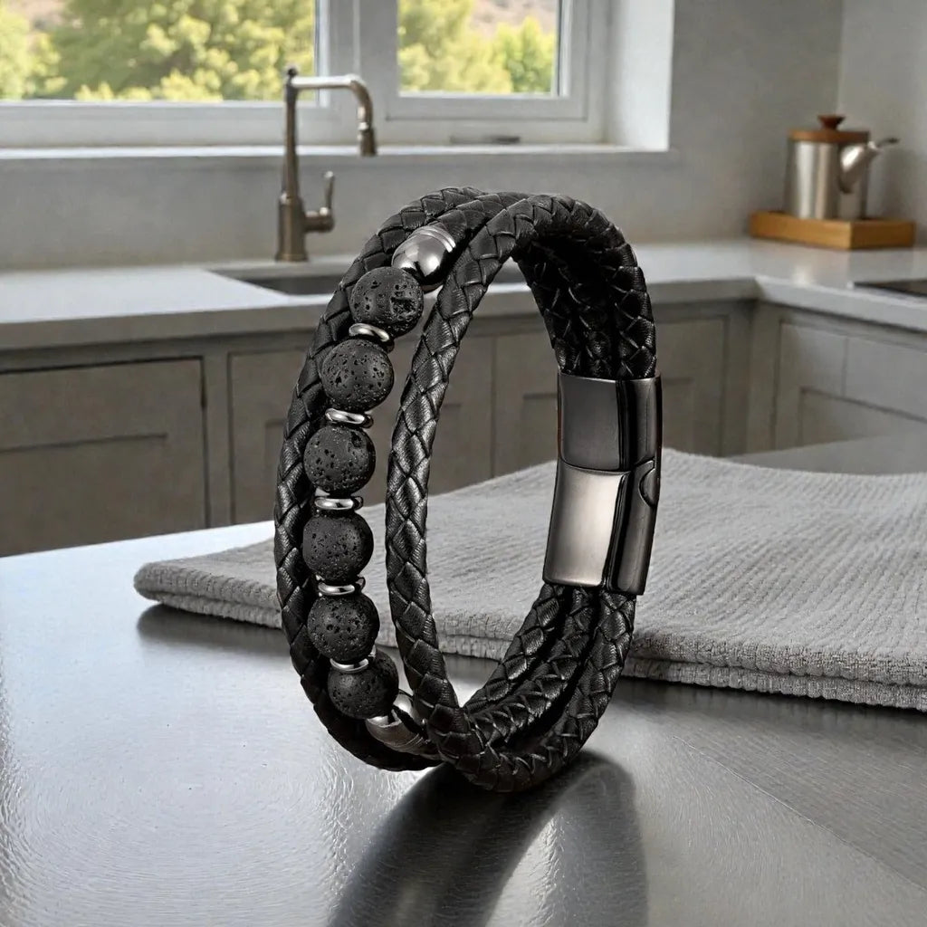 Le bracelet Vulcan en équilibre sur une table de cuisine satinée, mettant en relief l'aspect minéral des six perles volcaniques et le tressage complexe des trois lanières en cuir noir.