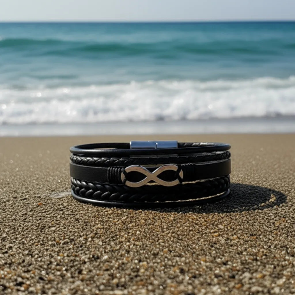 Le cuir noir multi-rangs à plat sur le sable. Le signe Infini en acier inoxydable argenté contraste avec le décor marin sous un ciel nuageux.