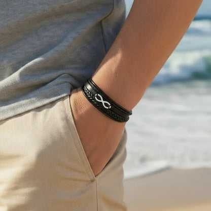 Gros plan. Le bracelet Infini en cuir noir au poignet d'un homme. Le signe Infini en acier ressort sur les vêtements beige et gris, main dans la poche.