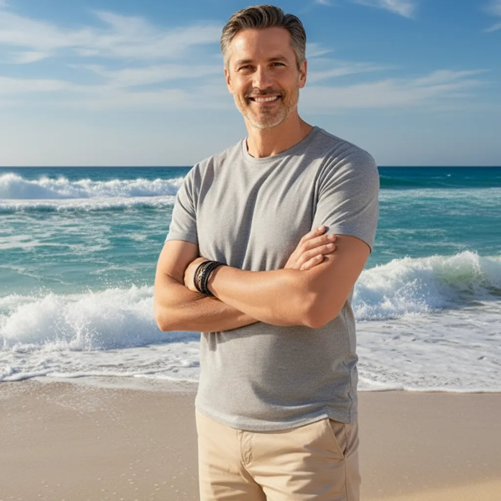 Jeune homme, bras croisés, attitude affirmée devant la mer agitée. Le bracelet Infini en cuir noir se distingue clairement au poignet gauche, sous un ciel bleu nuageux.