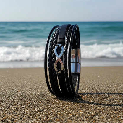 Vue latérale du bracelet Infini en cuir noir posé en équilibre sur le sable. Le fermoir magnétique en acier argenté est mis en évidence face à la mer agitée.