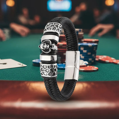 Scène de poker. Le bracelet Kaldor cuir tressé noir posé sur le bois, jetons et tapis vert en arrière-plan. Le symbole Pique et Tête de Mort et le maillage du risque captent la lumière de la salle de jeu.