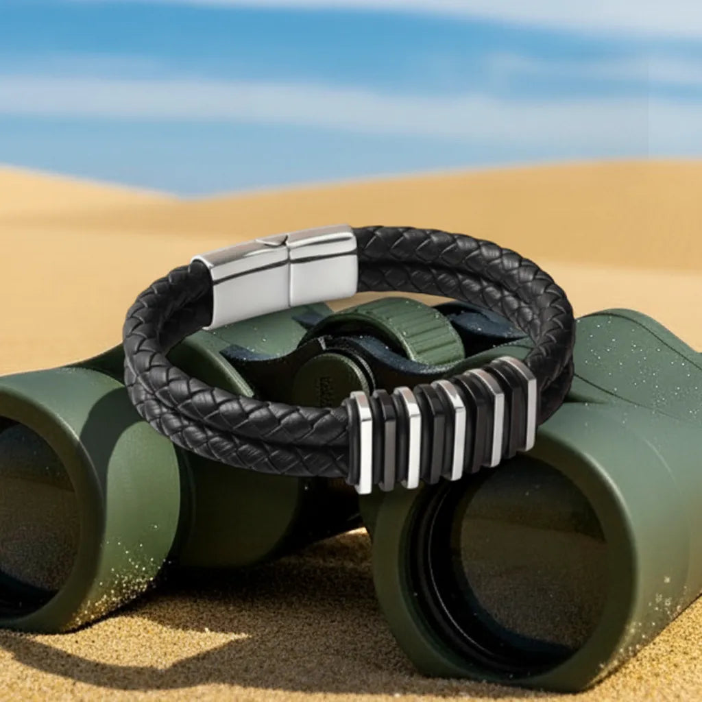 Vue technique. Le bracelet Link cuir tressé noir posé à plat sur les jumelles. La structure des segments contraste avec la texture du sable et des dunes.