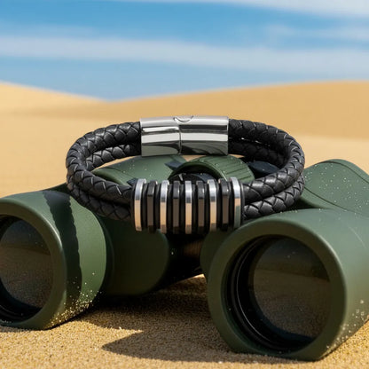 Détail de l'équipement. Vue de face du bracelet Link cuir noir tressé posé sur les jumelles. Le fermoir magnétique en acier inoxydable complète le design modulaire.