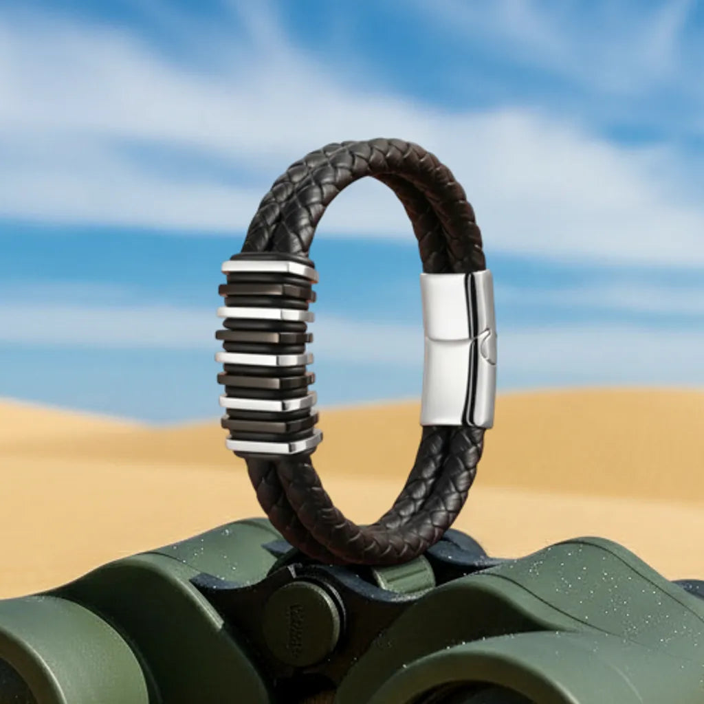 Ambiance exploration. Le bracelet Link cuir noir tressé dressé sur des jumelles militaires. Les segments modulaires en acier évoquent l'assemblage technique.