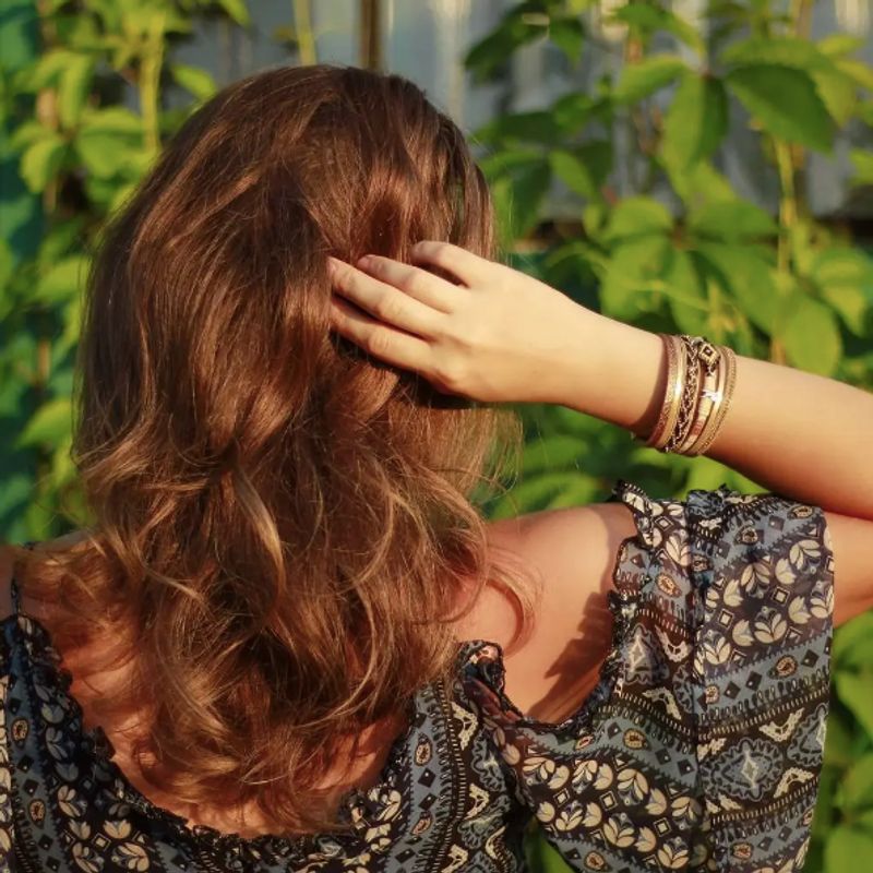 Bracelet mauvais œil, wrap multilanière en cuir PU beige et marron . Ce bracelet avec motif central représentant un mauvais œil en perles façon Miyuki, s'accorde avec une rivière de strass en cristal coloré et de fines chaînes en alliage de zinc. Présenté sur le poignet d'une femme rousse vue de dos. Élégante, elle est vêtue d'une robe bohème. Fermoir magnétique de couleur or.