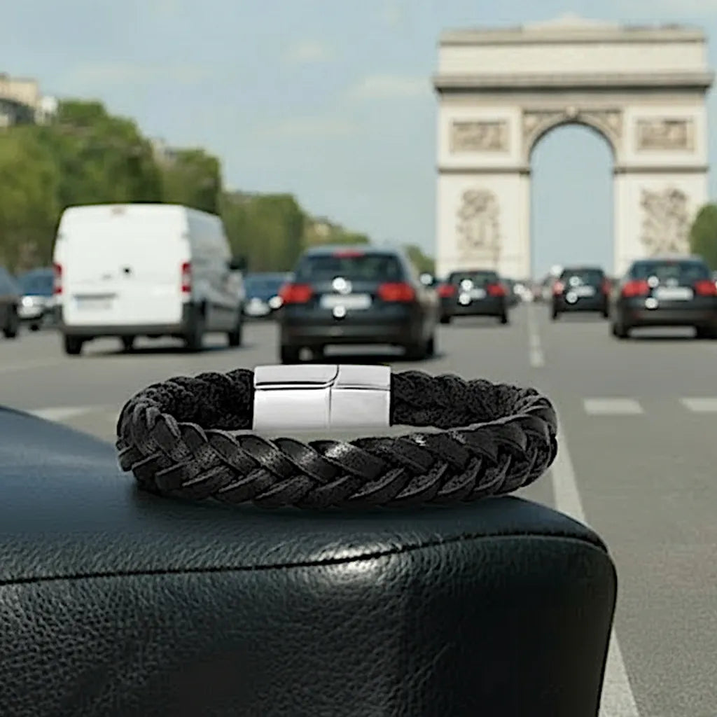 Plan de face. Le bracelet Nero Cuir Noir Tressé posé à plat sur la selle, avec l'Arc de Triomphe en toile de fond, insistant sur sa force tranquille.