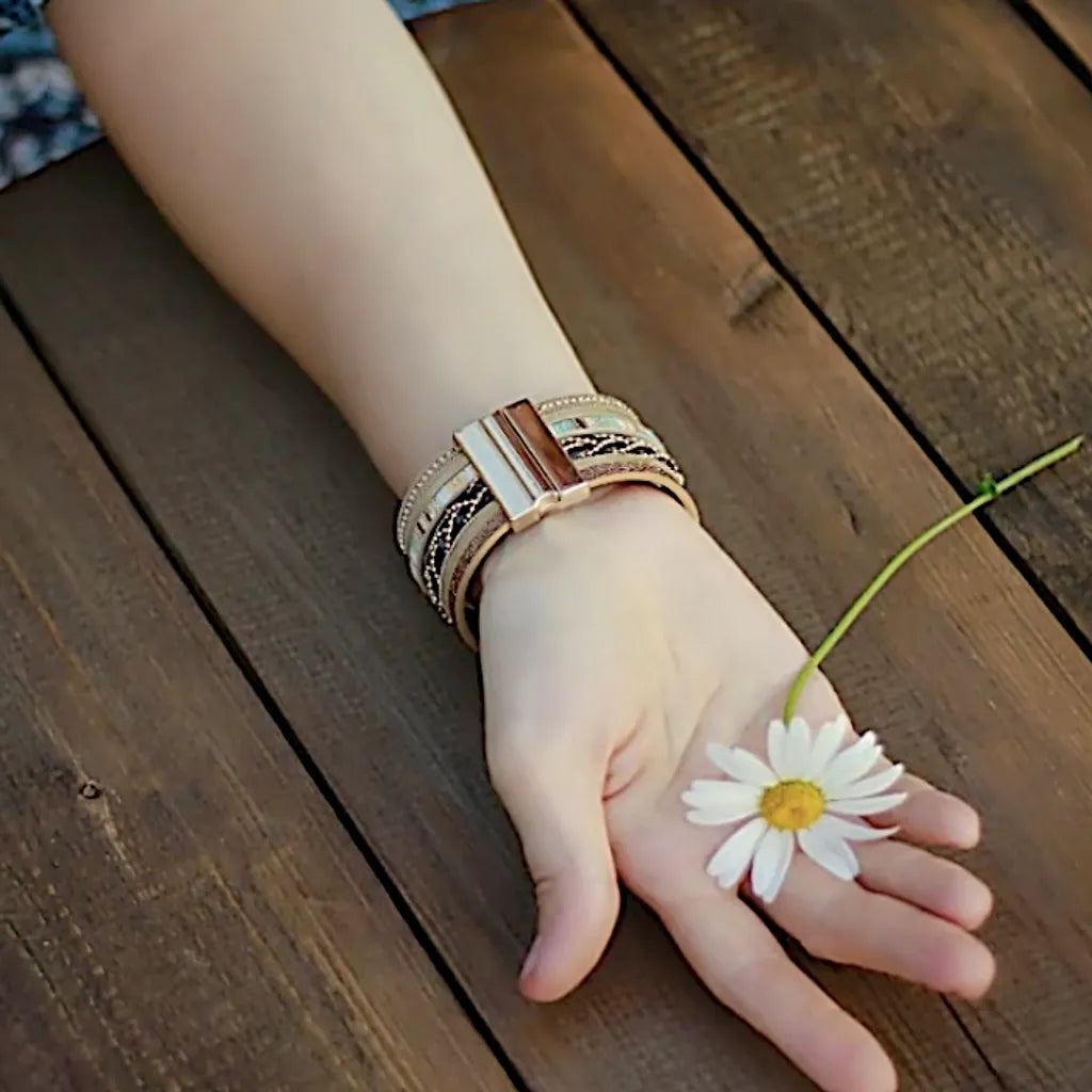Le bracelet Œil du Chaman multi-rangs, vu sur son fermoir doré magnétique, porté sur un poignet mis en valeur par une marguerite au creux de la main. Le style amérindien se marie à la nature.