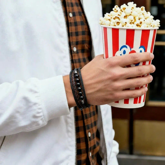 Focus sur le bracelet Oris au poignet, révélant la texture du cuir noir et l'éclat des perles agate noire. La main saisit un cornet de pop-corn devant l'entrée du cinéma, illustrant une élégance décontractée.