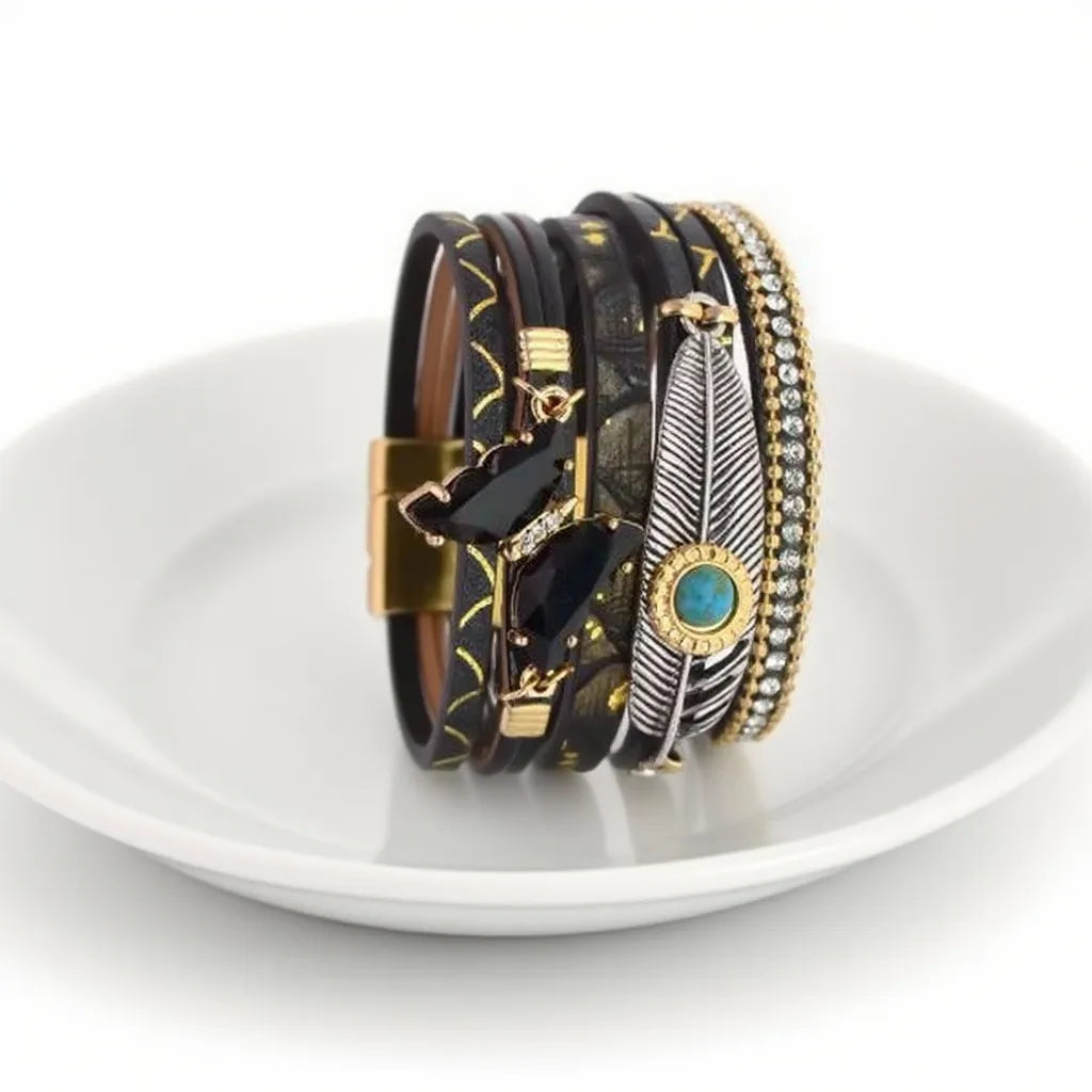 Vue de profil, au creux d'une assiette en porcelaine blanche du bracelet noir, détaillant le papillon en cristal, les chaînes fines, la plume Quetzal et le cabochon bleu cerclé doré.