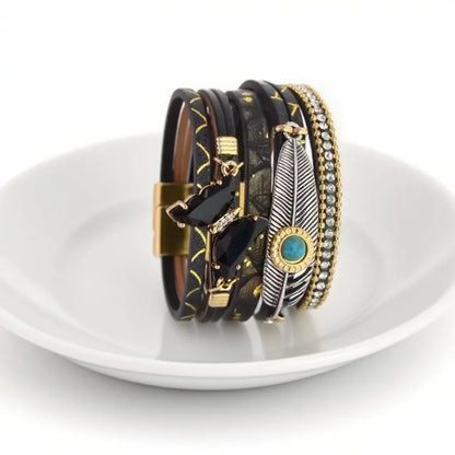 Vue de profil, au creux d'une assiette en porcelaine blanche du bracelet noir, détaillant le papillon en cristal, les chaînes fines, la plume Quetzal et le cabochon bleu cerclé doré.