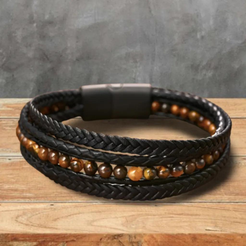 Bracelet multitours en cuir noir tressé, orné de perles œil de tigre jaune lumineux, posé à plat sur une table en bois naturel. L’arrière-plan gris nuancé met en valeur la chaleur des pierres naturelles et le contraste du cuir noir, pour un design sophistiqué et élégant.