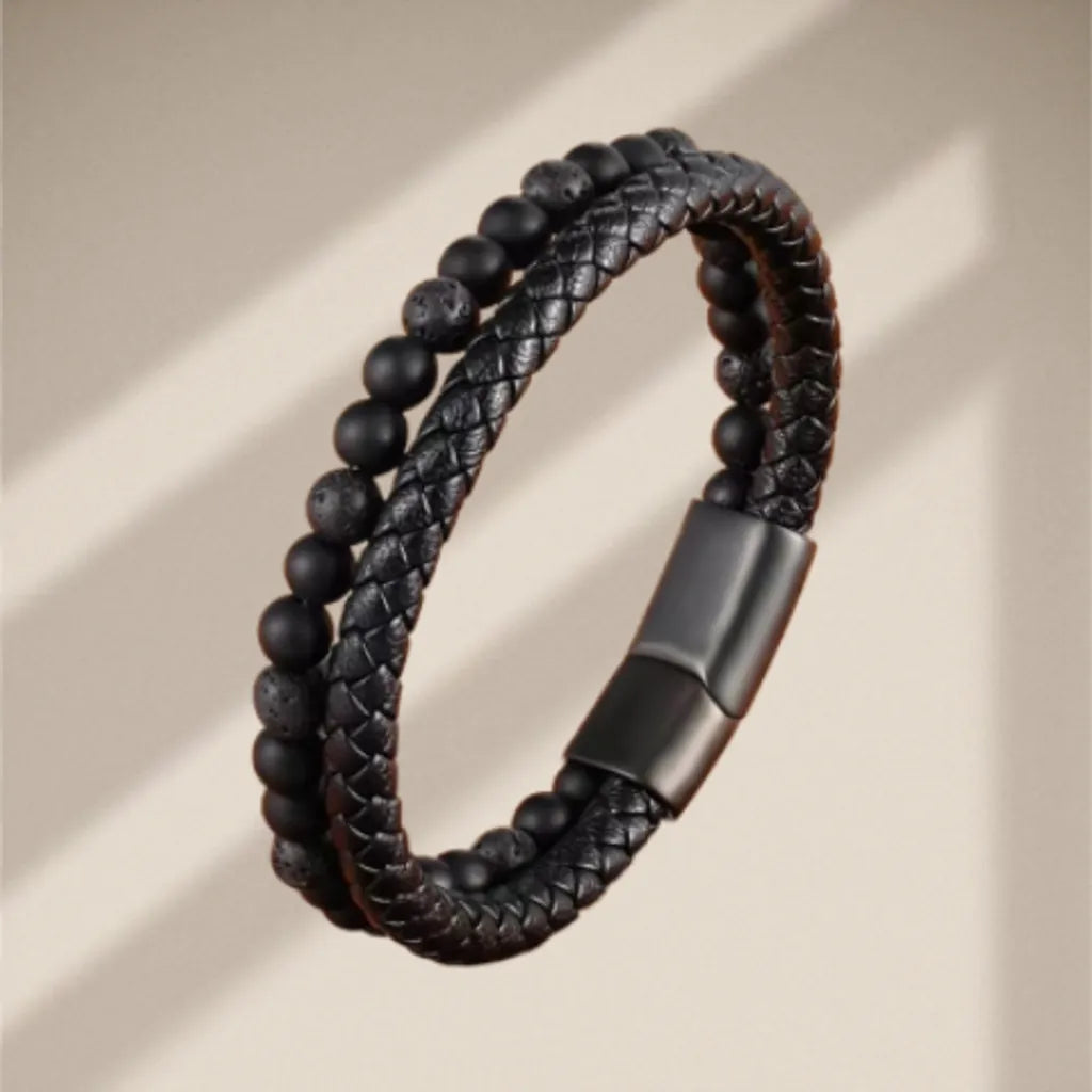 Bracelet en cuir tressé noir dressé, mettant en évidence ses perles naturelles de pierre de lave poreuse et d’onyx noir brillant. Le fond beige délicatement ombré souligne la verticalité et le design élégant du bracelet, parfait pour les amateurs d’accessoires à la mode.