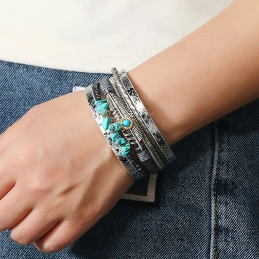 Bracelet plume bohème, ce wrap orné de pierre de couleur bleu turquoise est présenté autour d'un poignet d'une femme habillée en jean bleu. Multilanière en cuir PU gris, tresser ou lisse au motif peau de serpent.