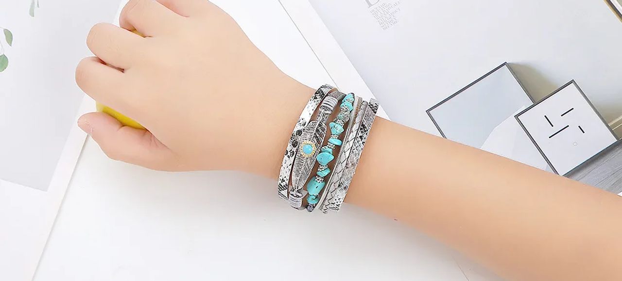 Bracelet plume bohème, ce wrap orné de pierre de couleur bleu turquoise est présenté autour d'un poignet d'une femme. Multilanière en cuir PU gris, tresser ou lisse au motif peau de serpent. Breloque Plume Bohème en alliage de zinc.