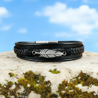 Vue de face du bracelet Plume Liberté multi-rangs en cuir noir, posé à plat sur un rocher, avec le ciel bleu en arrière-plan.