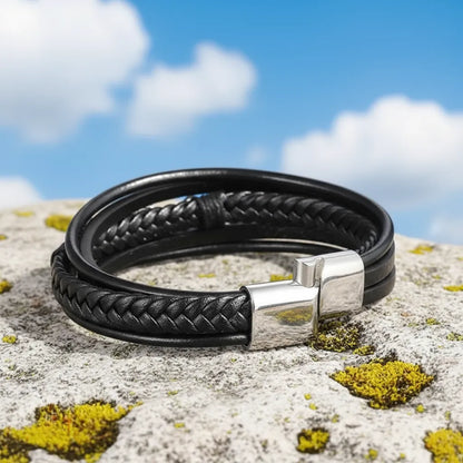 Vue de dos du bracelet en cuir noir. Gros plan sur le fermoir magnétique argenté et le multi-rangs, posés à plat sur un rocher, sous un ciel bleu nuageux.