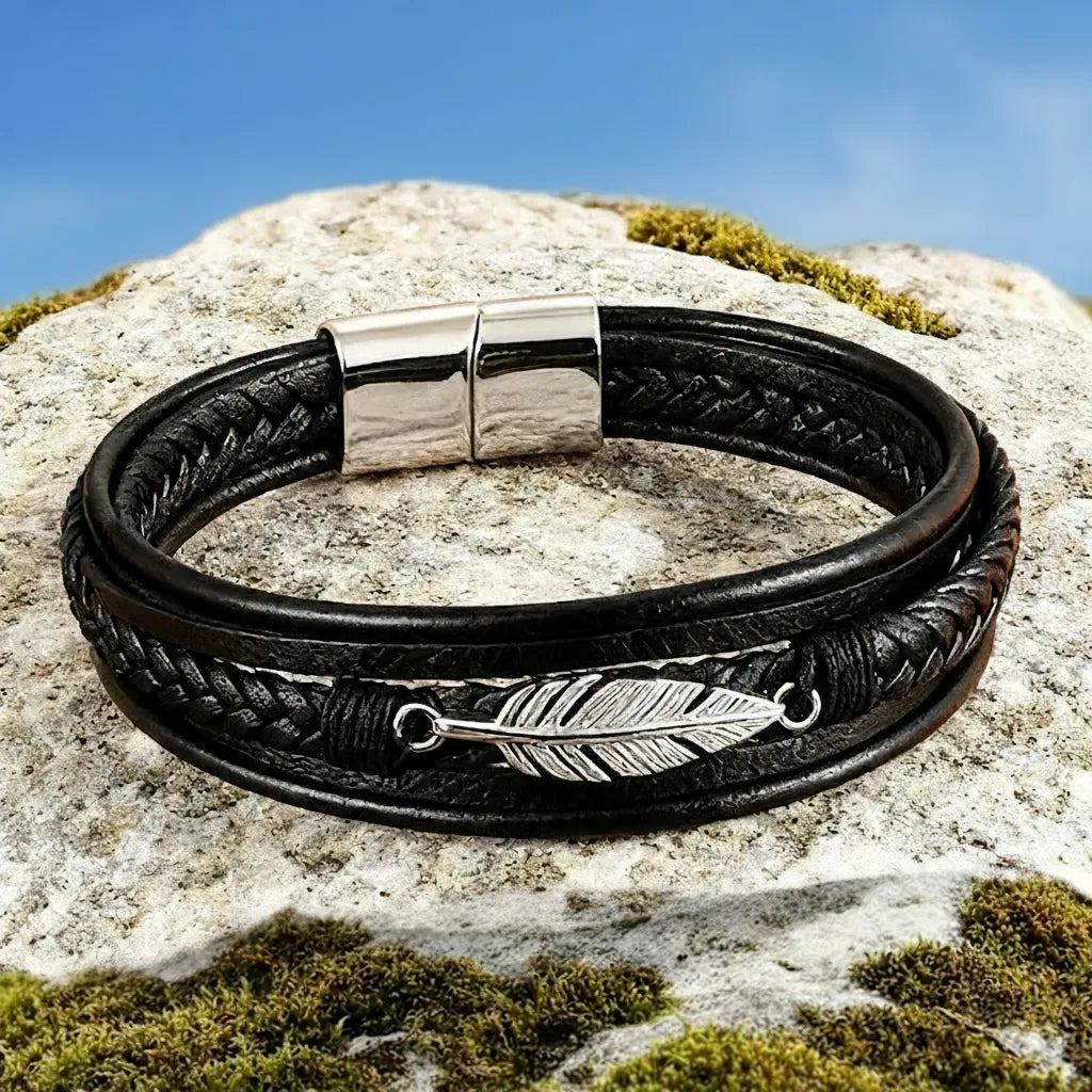 Bracelet en cuir noir multi-rangs posé à plat sur un rocher. Vue de haut de la plume Liberté et du fermoir en acier inoxydable argenté sur fond de ciel bleu.
