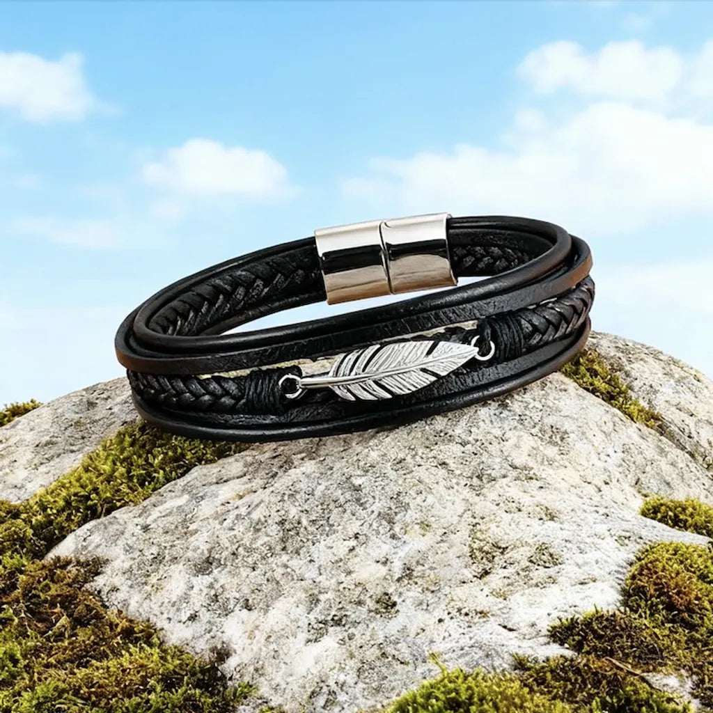 Bracelet multi-rangs en cuir noir et sa plume Liberté en acier argenté, posé à plat et légèrement penché sur un rocher sous un ciel bleu.
