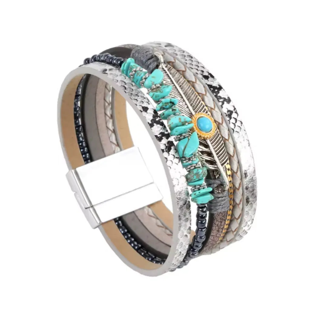 Bracelet Plume Quetzal argenté, vu gros plan, dressé ,du multi-rangs en cuir microfibre imprimé serpent gris, pierres turquoises et cabochon bleu cerclé doré, plume ajourée, rang perlé.