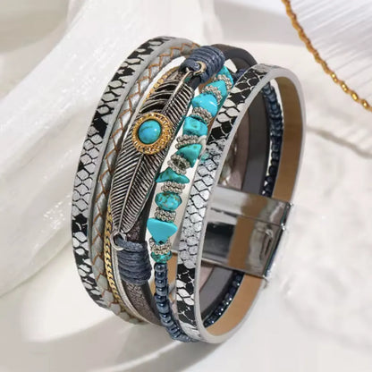 Bracelet Plume Quetzal argenté, cuir microfibre, cabochon bleu cerclé doré, plume ajourée et perles turquoise, fermoir argenté, prise lifestyle.