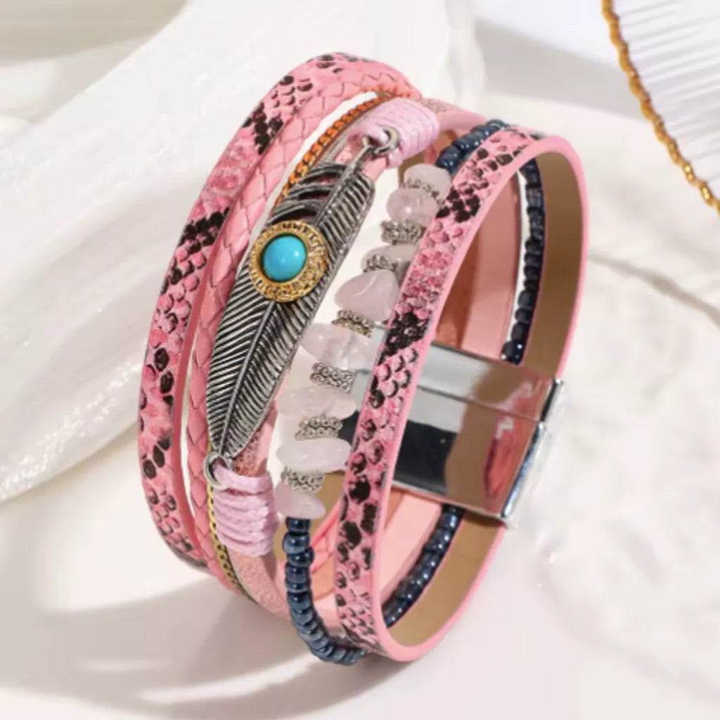 Bracelet en cuir microfibre rose multi-rangs, posé de côté. Il est orné d'une breloque plume ajourée, de perles noires et de pierres rose pâle alignées sur le même rang.