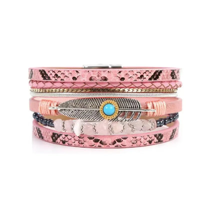 Bracelet en cuir microfibre rose délicatement courbé. Un rang de perles noires et de pierres rose pâle est aligné à côté des rangs lisses et d'une plume argentée à cabochon bleu.