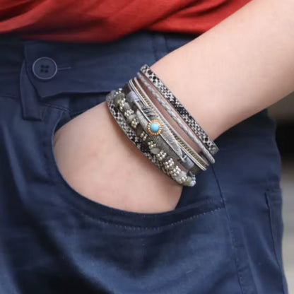 Bracelet multi-rangs en cuir microfibre gris porté sur un poignet, la main dans la poche avec breloque plume, perles noires et pierres grises assorties.