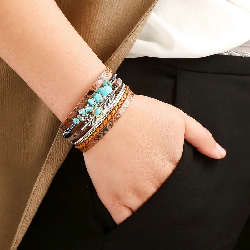 Bracelet plume bohème, ce wrap orné de pierre de couleur bleu turquoise est présenté autour d'un poignet d'une femme élégante habillée en pantalon noir. Wrap multilanière en cuir PU marron, tresser ou lisse au motif peau de serpent.