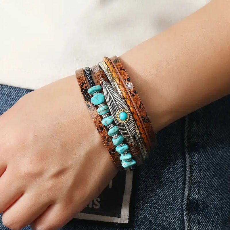 Bracelet plume quetzal, ce wrap orné de pierre de couleur bleu turquoise est présenté autour d'un poignet d'une femme habillée en jean bleu. Multilanière en cuir PU marron, tresser ou lisse au motif peau de serpent.