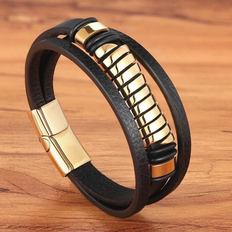 Bracelet spirale acier avec perle tube ajourée, dorée et multilanère en cuir noir. Fermoir magnétique en acier inoxydable de couleur or. Ce wrap est présenté fermé, dressé sur un plan de travail en bois clair.