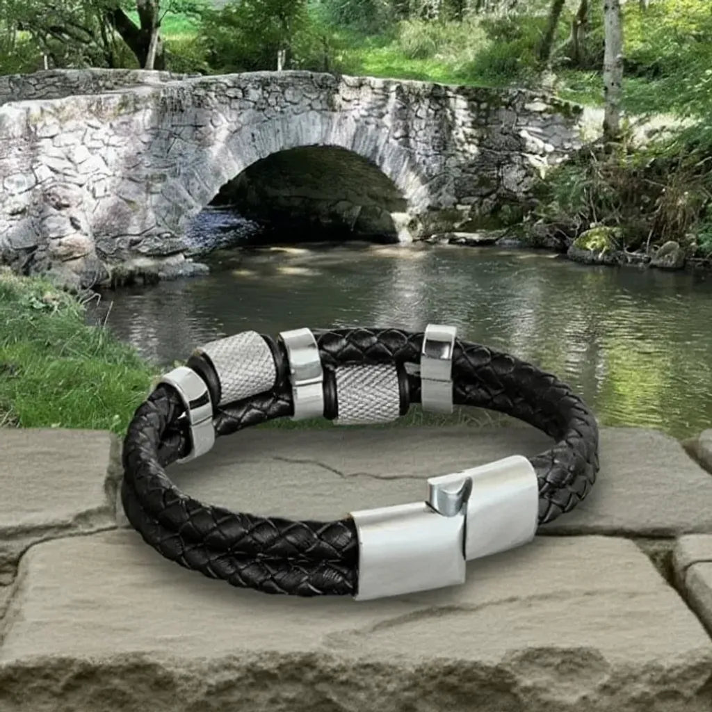 Détail technique. Gros plan sur le fermoir magnétique acier inoxydable entrouvert. Le bracelet Talon cuir tressé posé à plat, ses cylindres moletés dominant la rivière du Tarn.