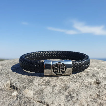 Vue de face du bracelet Templiers en cuir noir tressé avec fermoir magnétique Croix Pattée, posé sur un rocher sous un ciel bleu.