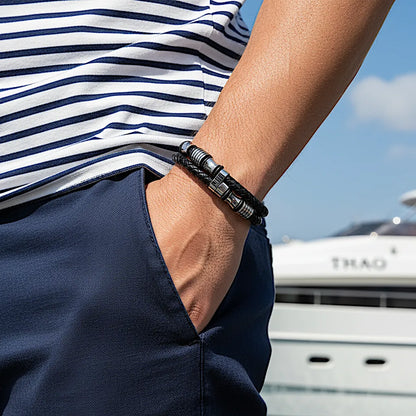 Gros plan. Le bracelet Thao affirme l'allure nautique. La double lanière en cuir noir tressé et les perles sont mises en valeur devant le yacht.