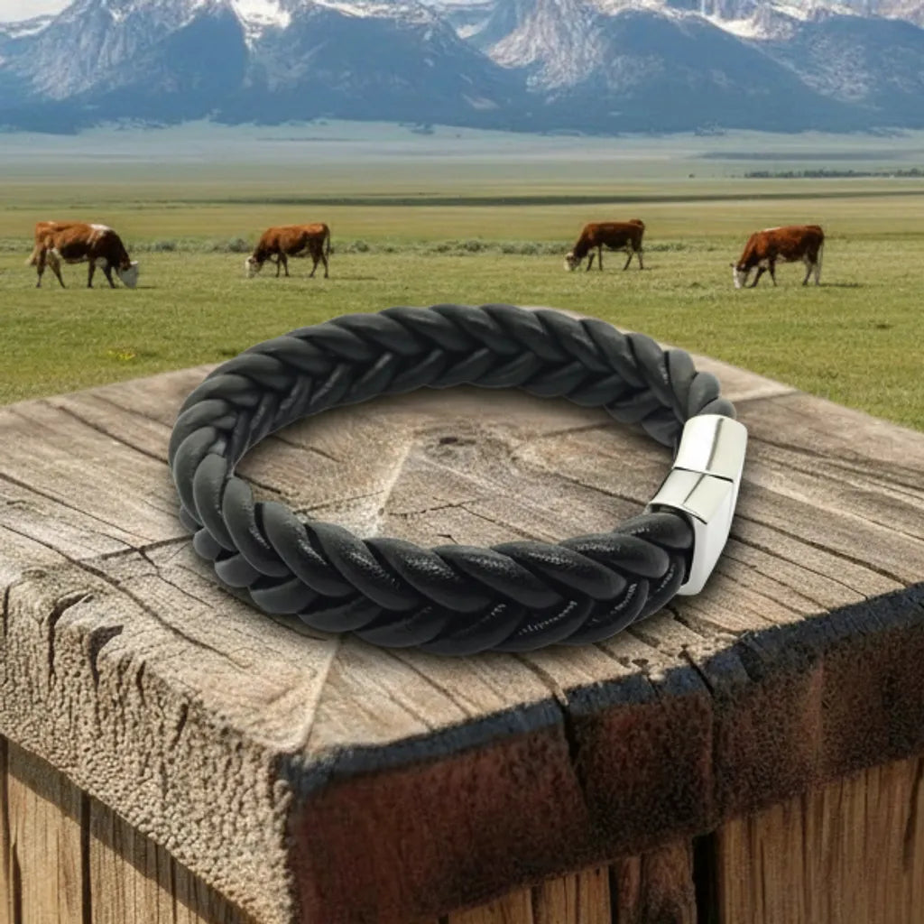 Gros plan paysage. Le bracelet Torc cuir souple noir, avec son tressage visible, repose sur le pilier en bois. Le volume du cuir tressé s'intègre au charme de la campagne.