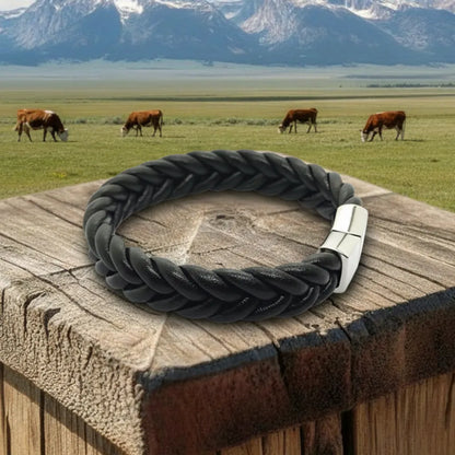 Gros plan paysage. Le bracelet Torc cuir souple noir, avec son tressage visible, repose sur le pilier en bois. Le volume du cuir tressé s'intègre au charme de la campagne.