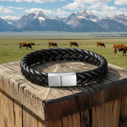 Vue en extérieur. Le bracelet Torc posé à plat sur le bois, son volume artisanal et son fermoir en acier inoxydable en premier plan, face à la prairie et aux vaches.