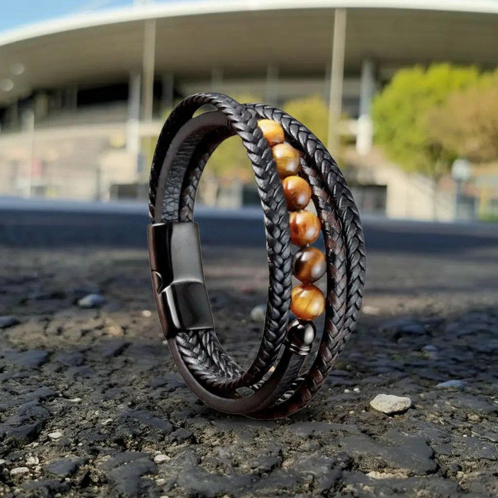 Composition dynamique sous un ciel azur, valorisant la lanière plate texturée qui soutient les six billes de perles oeil de tigre. L'environnement urbain boisé encadre cet assemblage technique de cuir tressé et de minéraux.