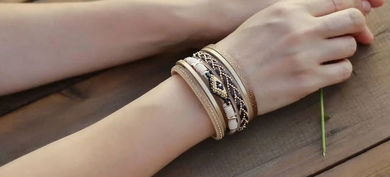 Bracelet wrap avec motif mauvais œil en perles Miyuki. Multilanière bohème en cuir PU de couleur beige et marron portée autour d'un poignet d'une jeune femme posé sur une table rustique. 