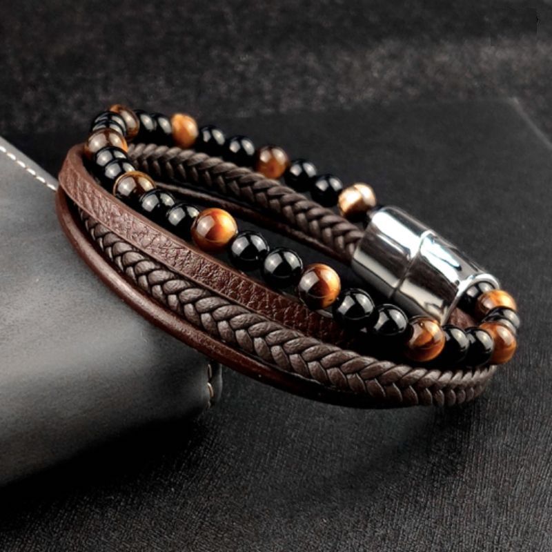 Bracelet de pierres naturelles agate noire et œil de tigre jaune pour homme, son fermoir magnétique en acier inoxydable est argenté – posé et incliné sur le coin d'un livre à la reliure en cuir gris  – wrap cuir coloris marron.