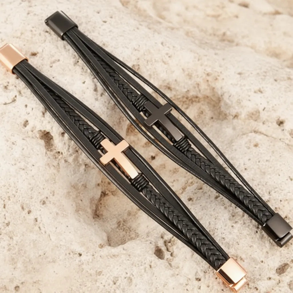 Vue de haut, à plat, de deux bracelets multi-rangs en cuir noir, doré et noir, avec croix latine et fermoir ouvert, sur un rocher.