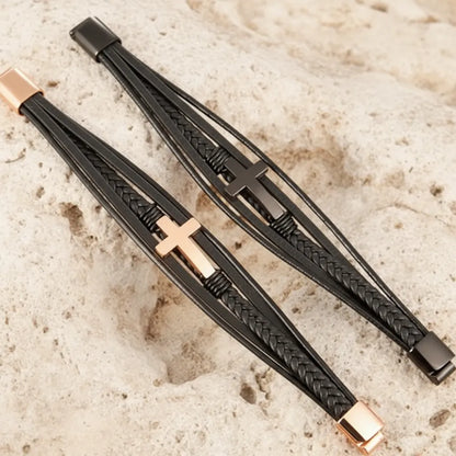 Vue de haut, à plat, de deux bracelets multi-rangs en cuir noir, doré et noir, avec croix latine et fermoir ouvert, sur un rocher.