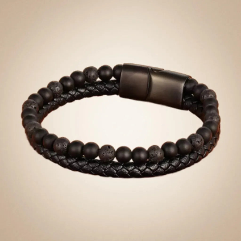 Bracelet en cuir tressé noir orné de perles de pierre de lave texturées et de perles d’onyx noir brillant. Le fermoir magnétique noir en métal mat apporte une touche moderne et élégante. L’accessoire repose sur un fond beige doux avec un subtil effet d’ombre, mettant en avant son superbe design et ses matériaux naturels, idéal pour une allure masculine et intemporelle.