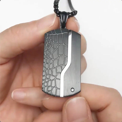 Gros plan du collier Oblong noir avec pendentif à plaque au motif terre aride en acier inoxydable, présenté entre les doigts de deux mains. Présence d'un zircone sur le côté gauche inférieur - coloris noir.