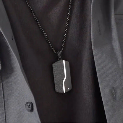Gros plan du collier Oblong noir porté, avec pendentif moderne en acier porté par un homme vêtu de noir. Présence d'un zircone sur le côté gauche inférieur - coloris acier noir.