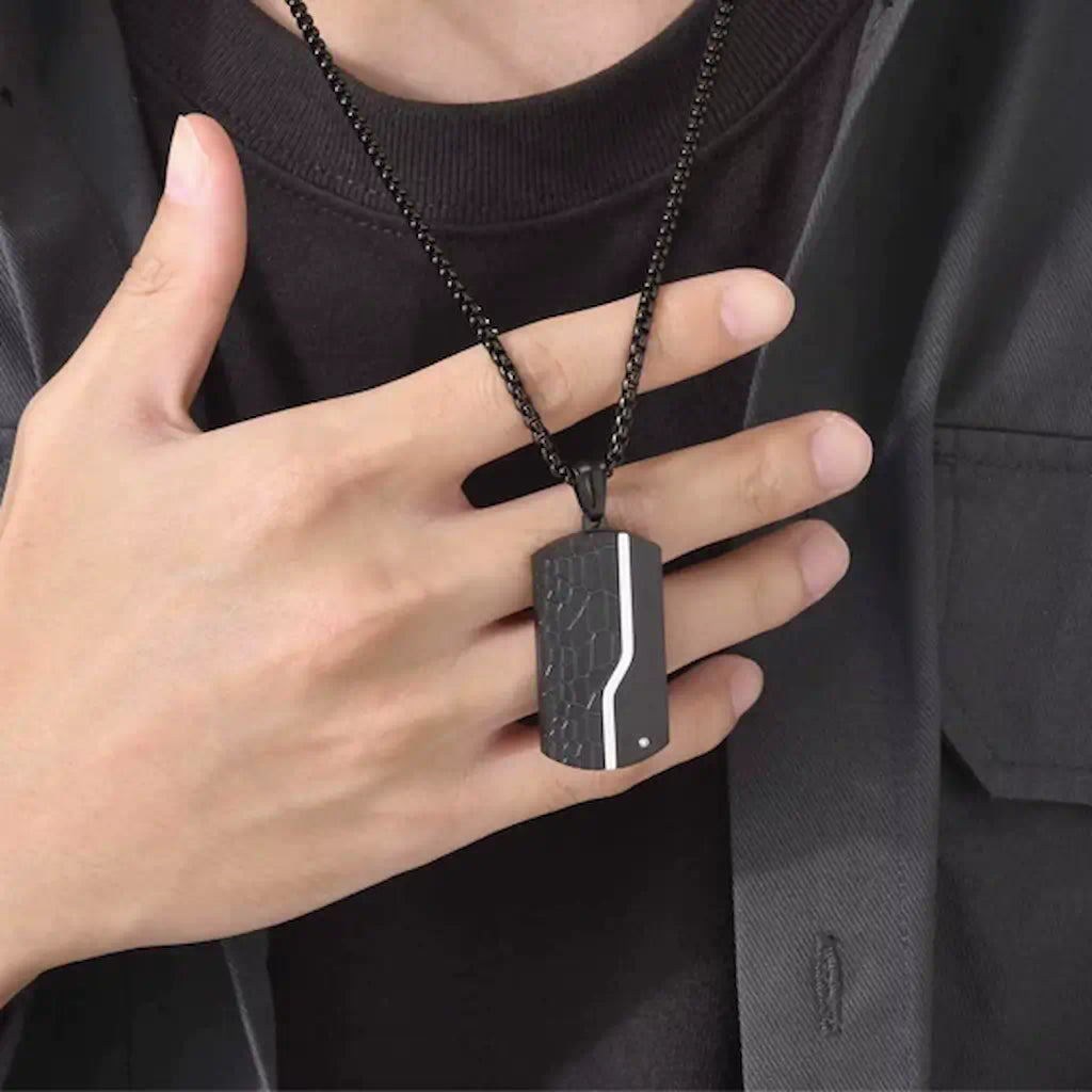 Gros plan du collier Oblong noir avec pendentif à plaque en acier porté par un homme vêtu de noir. Présence d'un zircone sur le côté gauche inférieur - coloris noir.
