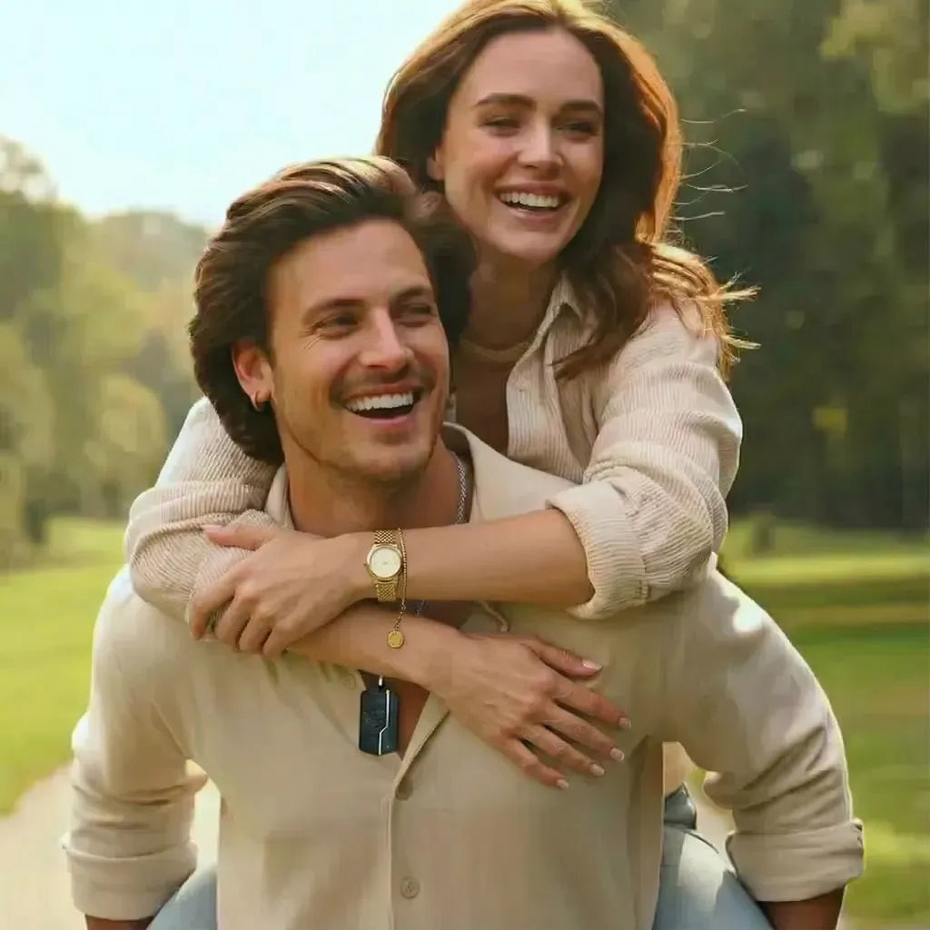 Couple heureux, l'homme porte sur son dos une femme radieuse, la scène est printanière sur un chemin ensoleillé. Chemises beiges, collier pendentif homme, collier et bracelet montre maille milanaise dorés. Joie et bijoux minimalistes.